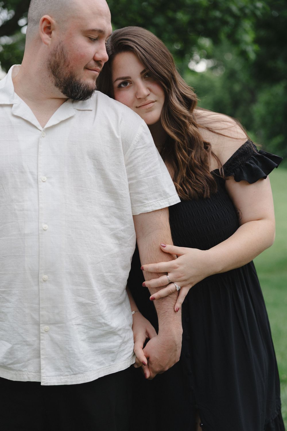 Couple embracing outdoors, woman in black off-shoulder dress with engagement ring, man in white shirt, green trees background.