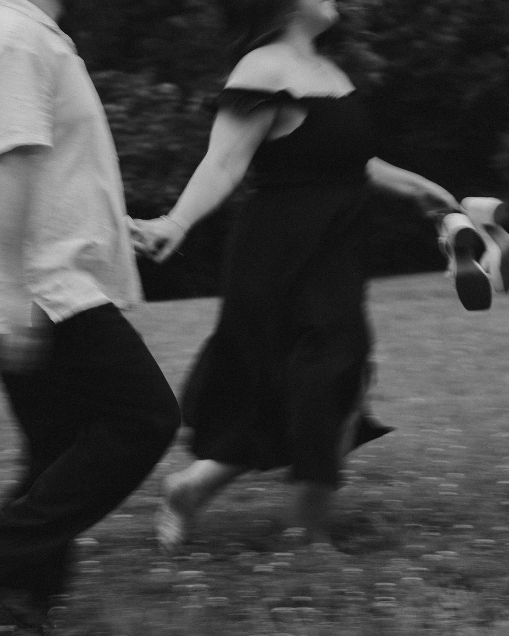 Black and white photo of a couple holding hands and running through a grassy field, woman in off-shoulder dress.