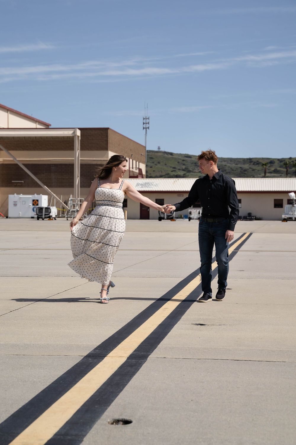 A couple dances playfully on an empty tarmac with mountains in the background.