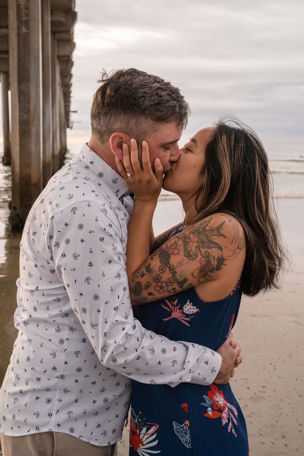 A romantic couple shares a kiss under a pier by the ocean.