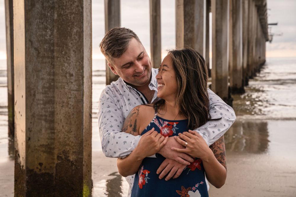 A couple laughs together while embracing under a pier by the beach.