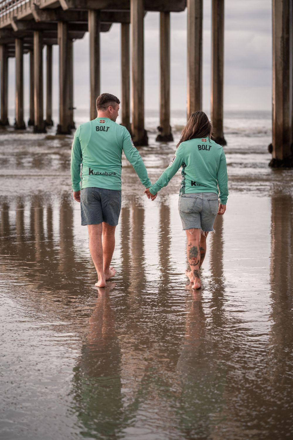 A couple in matching green sweatshirts walks hand in hand under a pier.