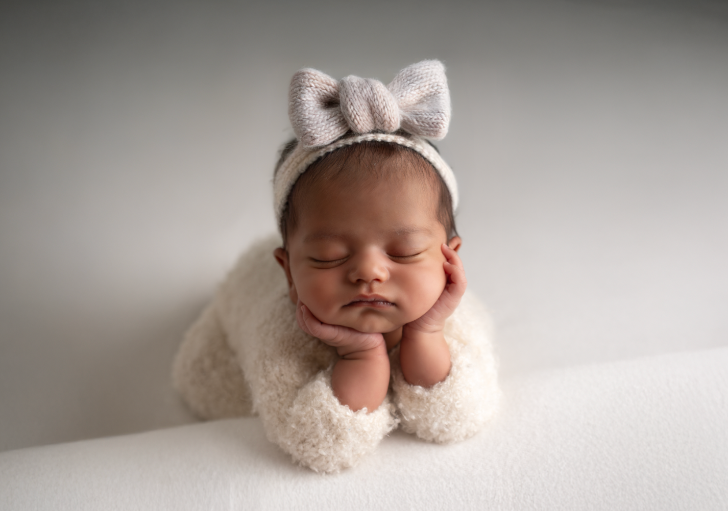 Sleeping newborn baby wearing a knit bow headband and fuzzy white outfit, posed with hands under chin.