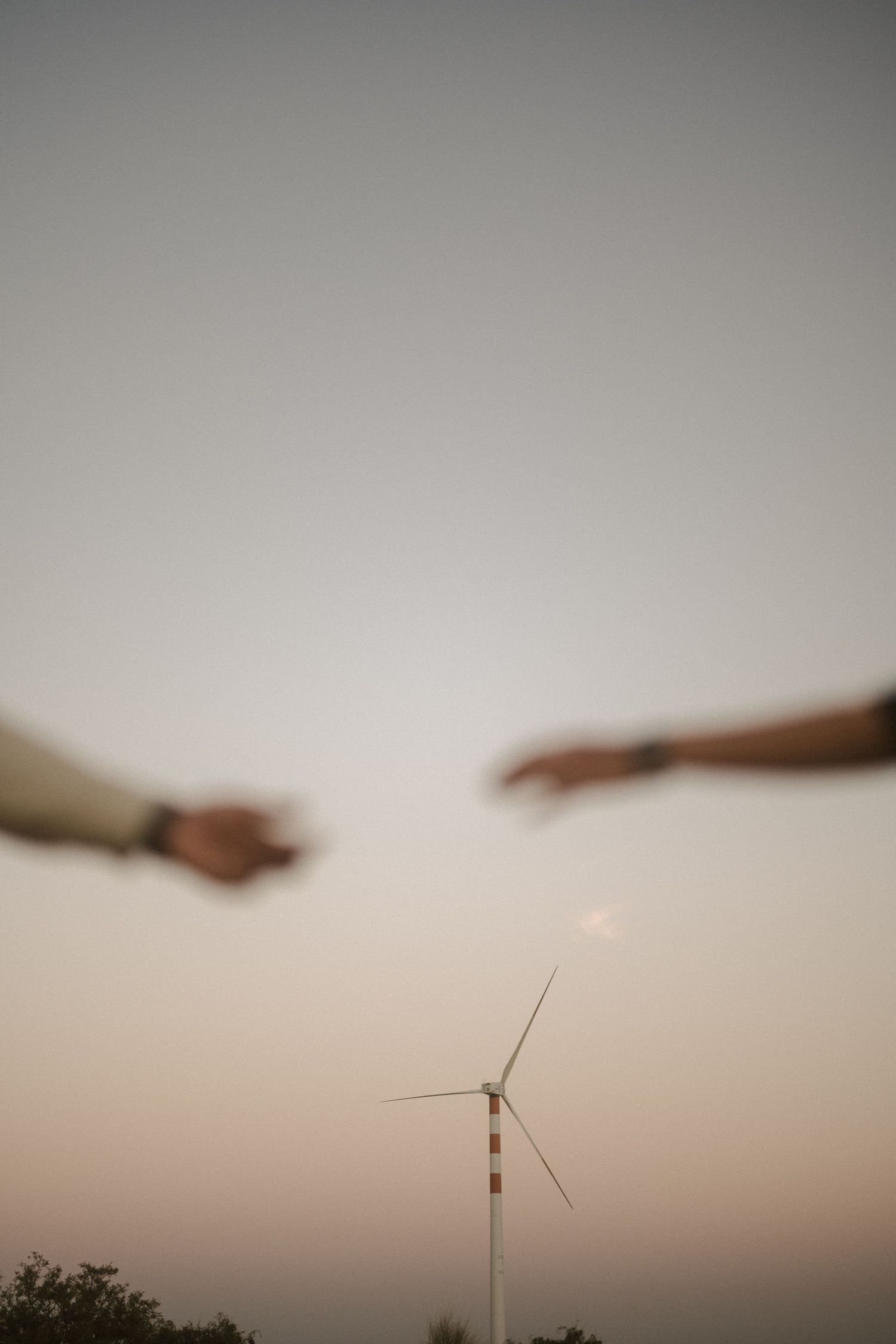 Two hands reaching towards a wind turbine silhouetted against a sunset sky.