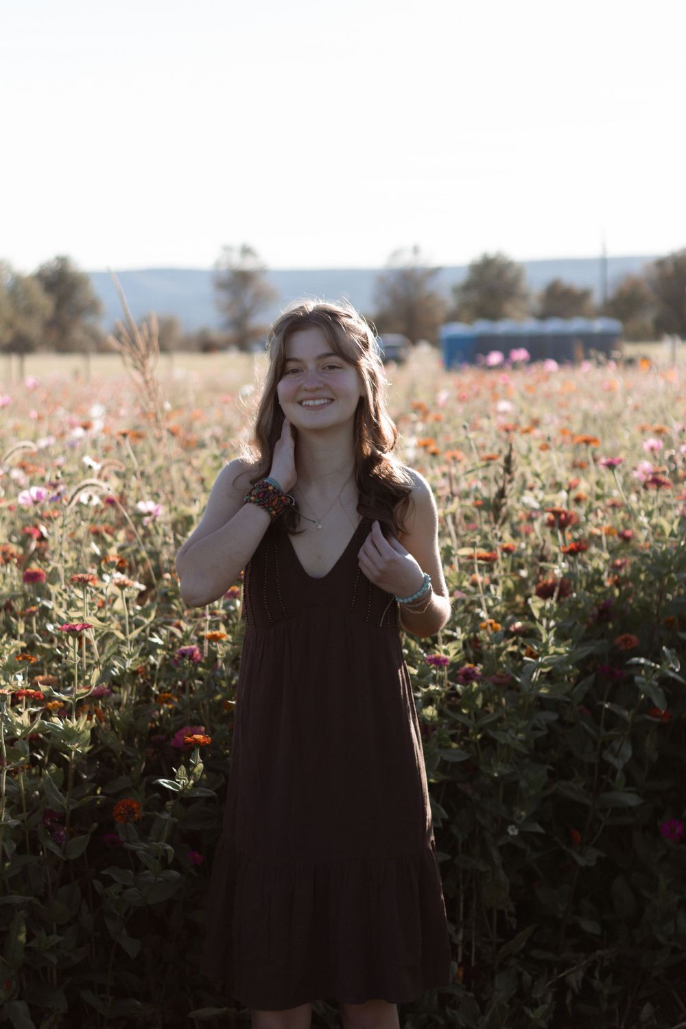 Woman in brown dress poses playfully in front of blooming flower field at sunset.