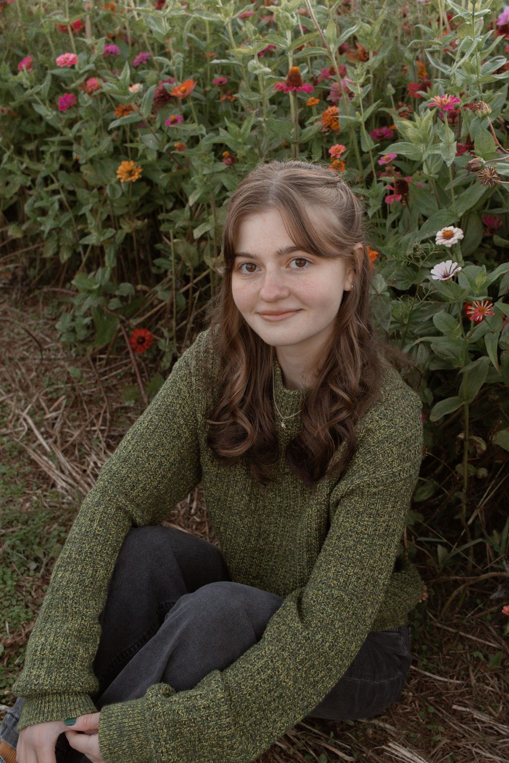 Person in green sweater poses among colorful flowers in a garden setting.