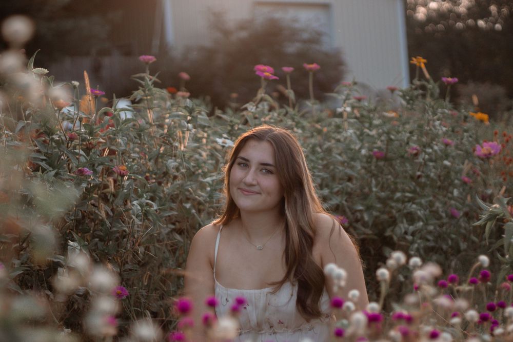 A white sundress-clad figure poses in a dreamy garden filled with pink and white wildflowers at sunset.
