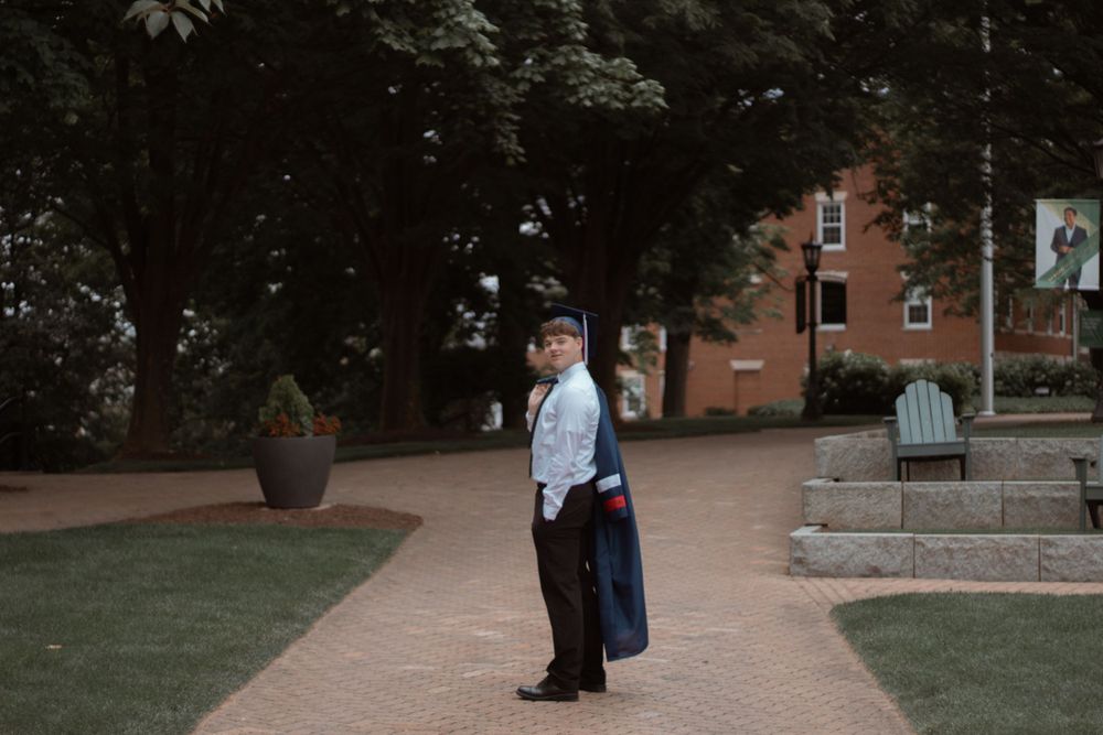 A graduate stands on a brick pathway in front of a building wearing traditional commencement attire.