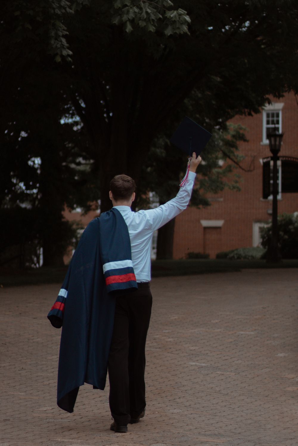 Graduate in cap and gown waves goodbye in front of a brick university building.