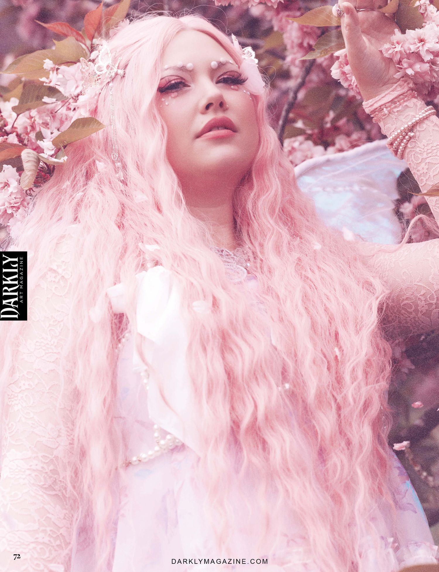 A woman dressed as a fairy with pink hair and wings, standing among pink blossoms. The image is from Darkly Art Magazine