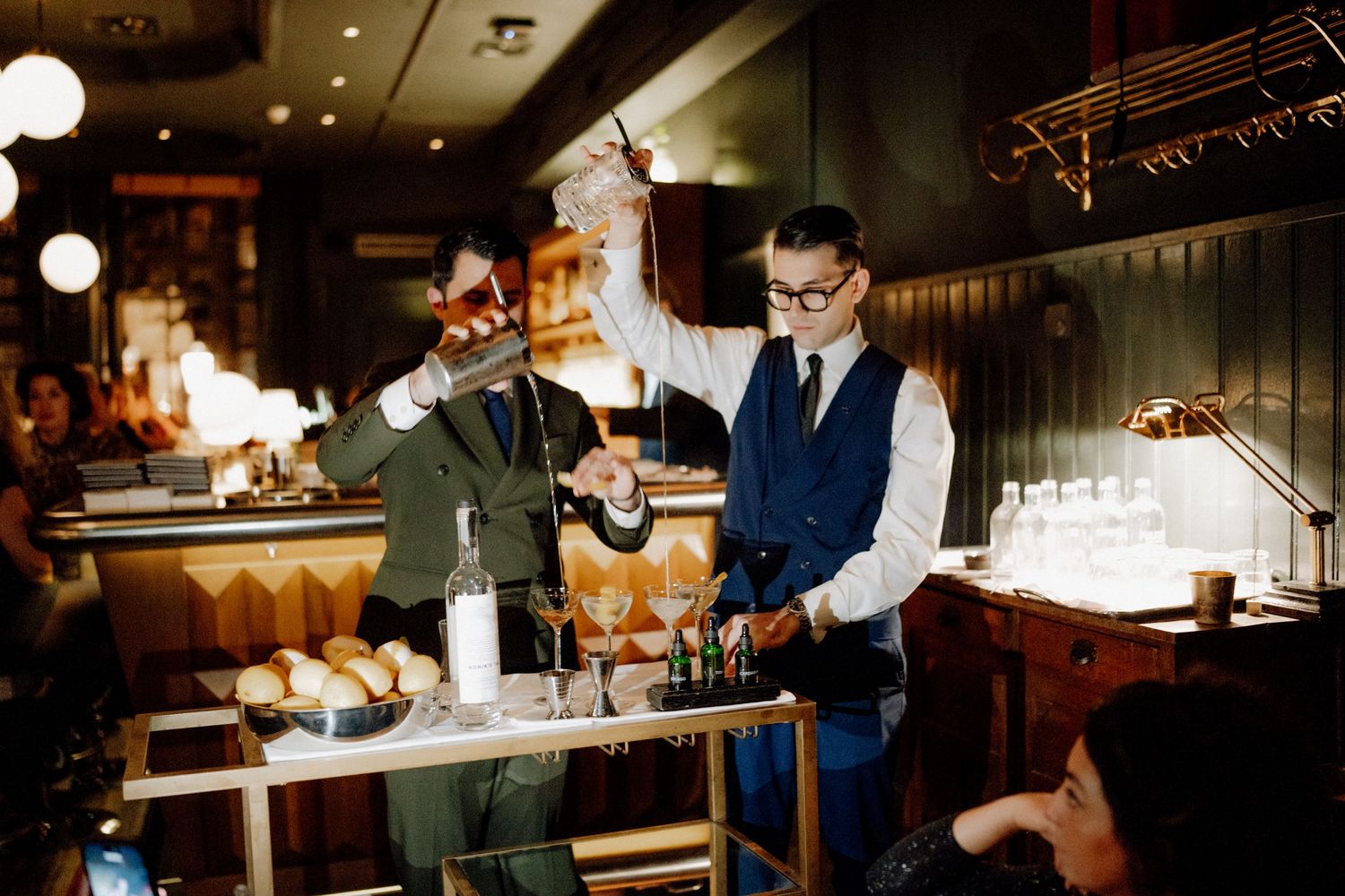 Two bartenders working behind a dimly lit bar counter with flames and bottles visible in the background.