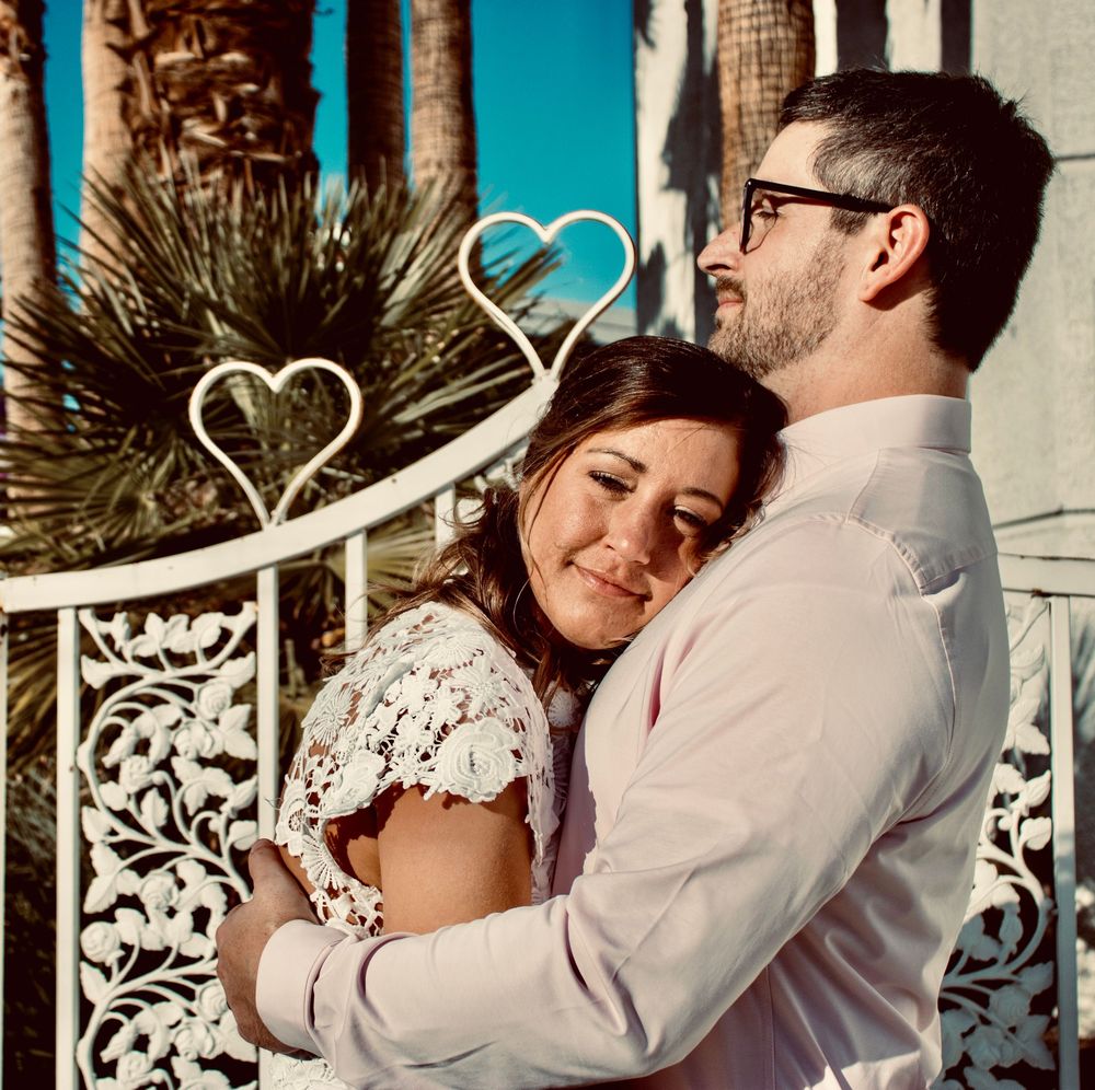 Couple embracing romantically outdoors with heart-shaped decorations, palm trees, and ornate white ironwork in background.