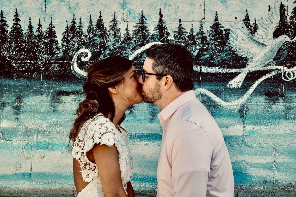 Couple sharing a romantic kiss in front of a colorful mural with trees and abstract art.