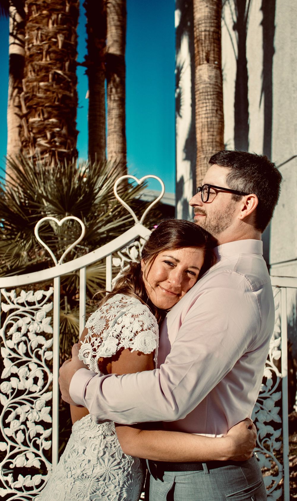 A couple embraces tenderly in wedding attire near heart-shaped decorations and palm trees under a bright blue sky.