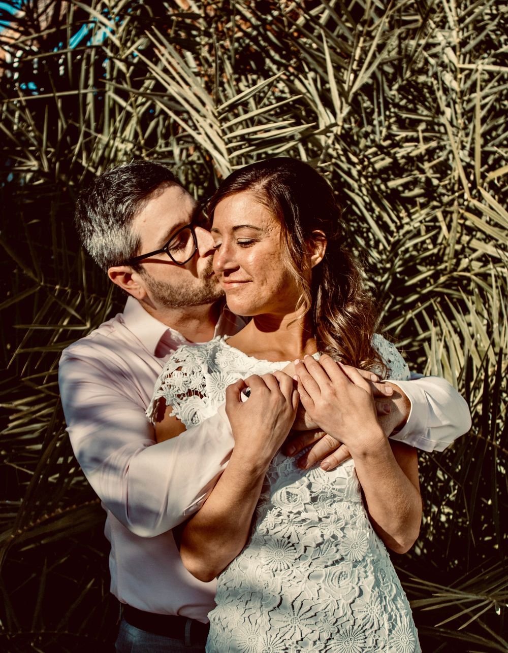 A couple embraces tenderly outdoors, man kissing woman's cheek near tropical plants in warm sunlight.