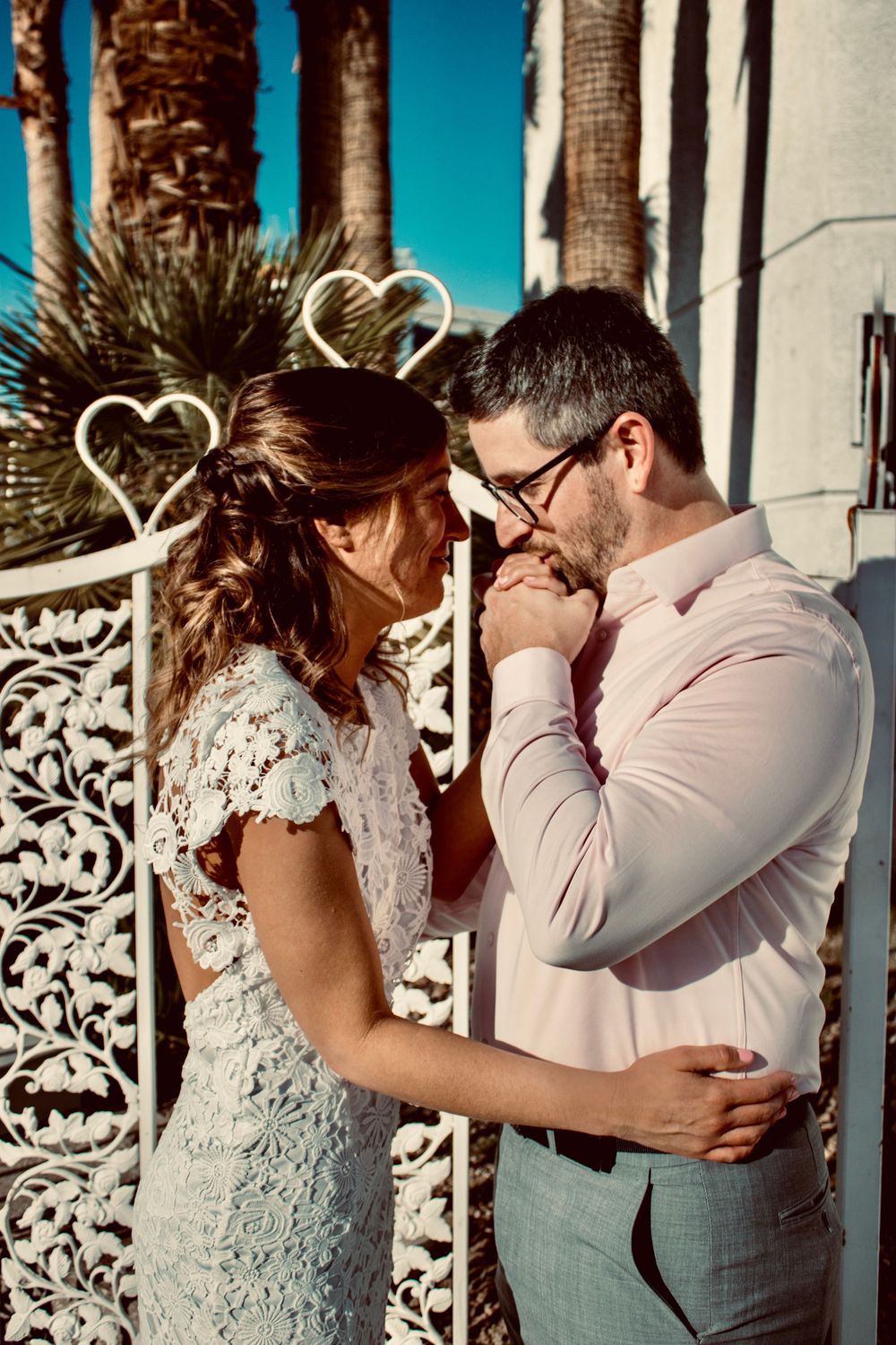 Couple in wedding attire sharing a romantic moment by ornate white gate with heart decorations and palm trees.