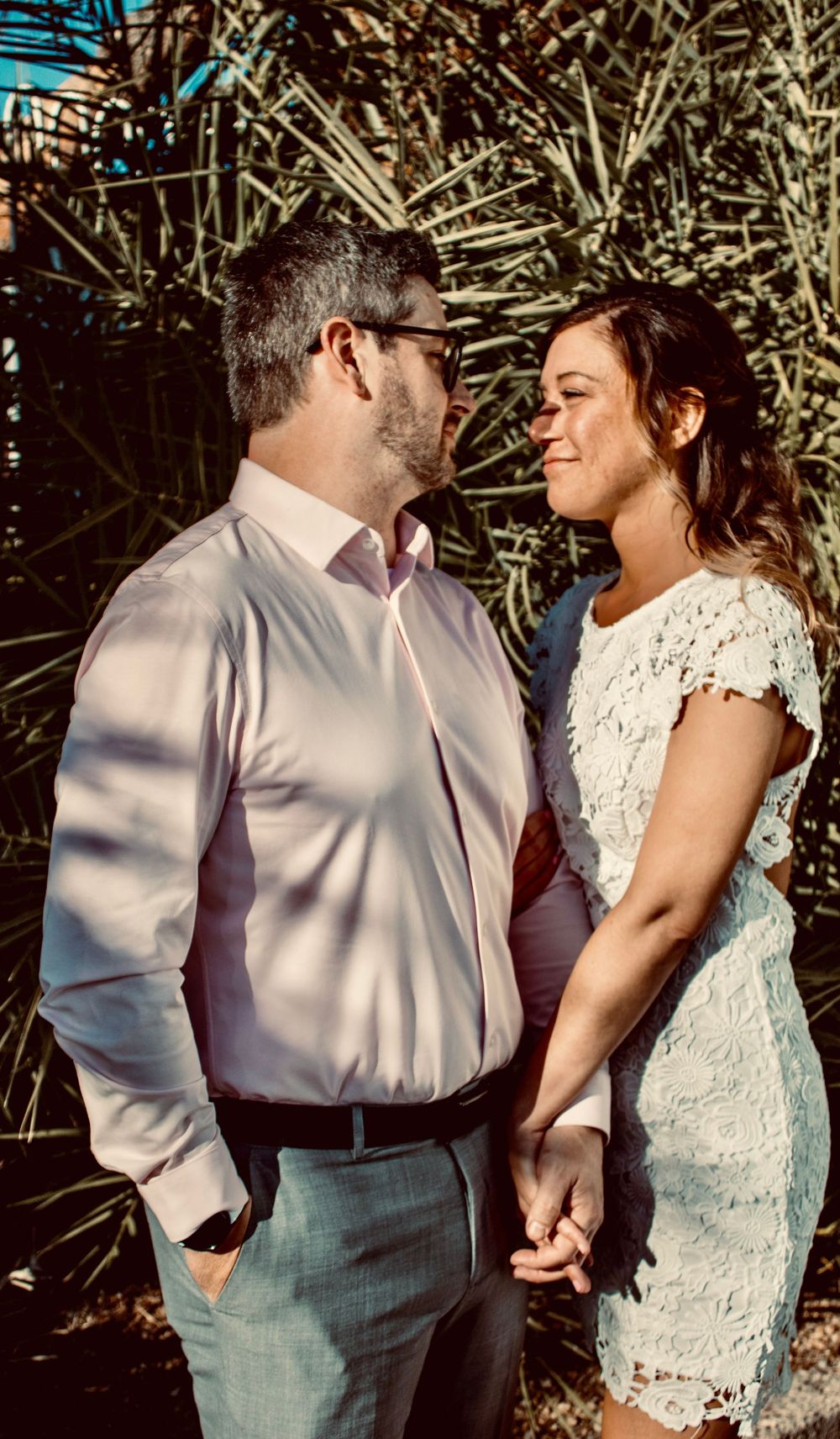 Couple gazing lovingly at each other holding hands outdoors near tropical plants in wedding attire.