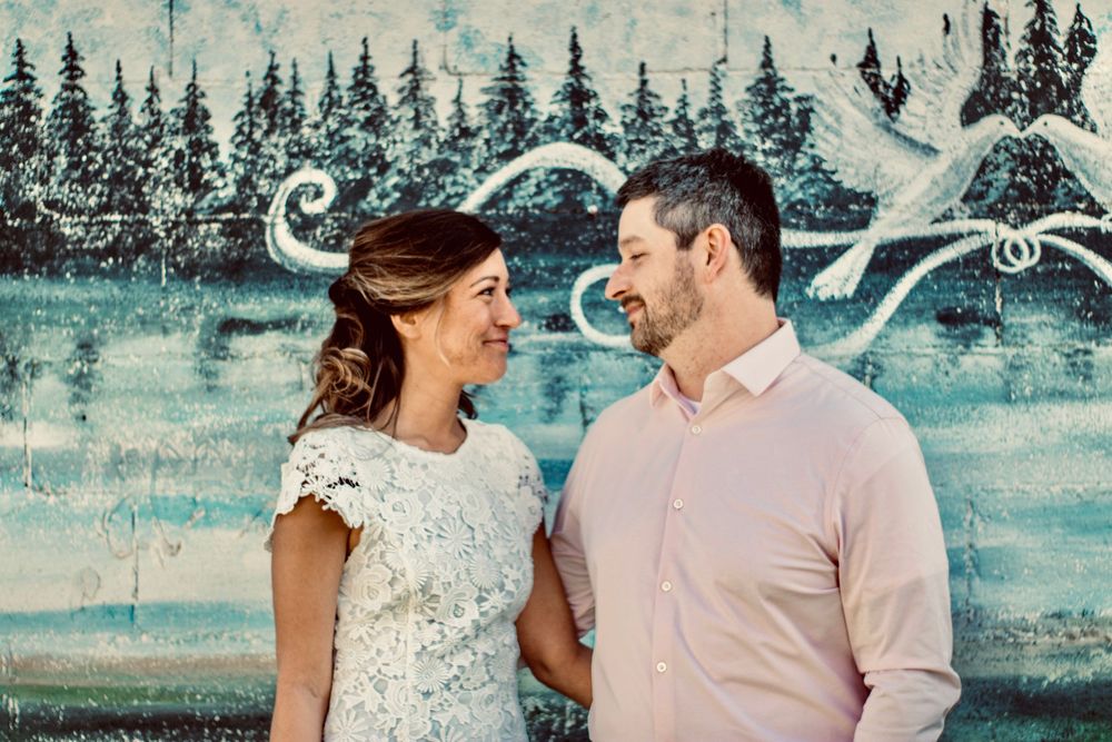 Couple smiling at each other in front of a colorful forest mural with a white bird painting.