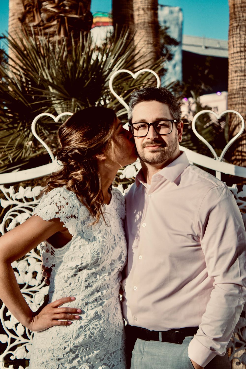 Bride in white lace dress kisses groom in pink shirt on cheek near heart-shaped decor in Las Vegas.