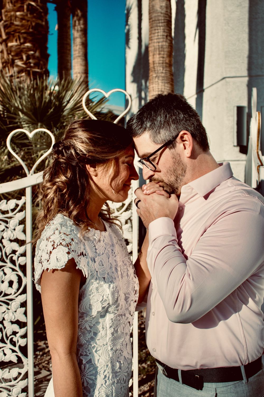 Groom in pink shirt gently kisses bride's hand near ornate white heart gate at Las Vegas chapel.