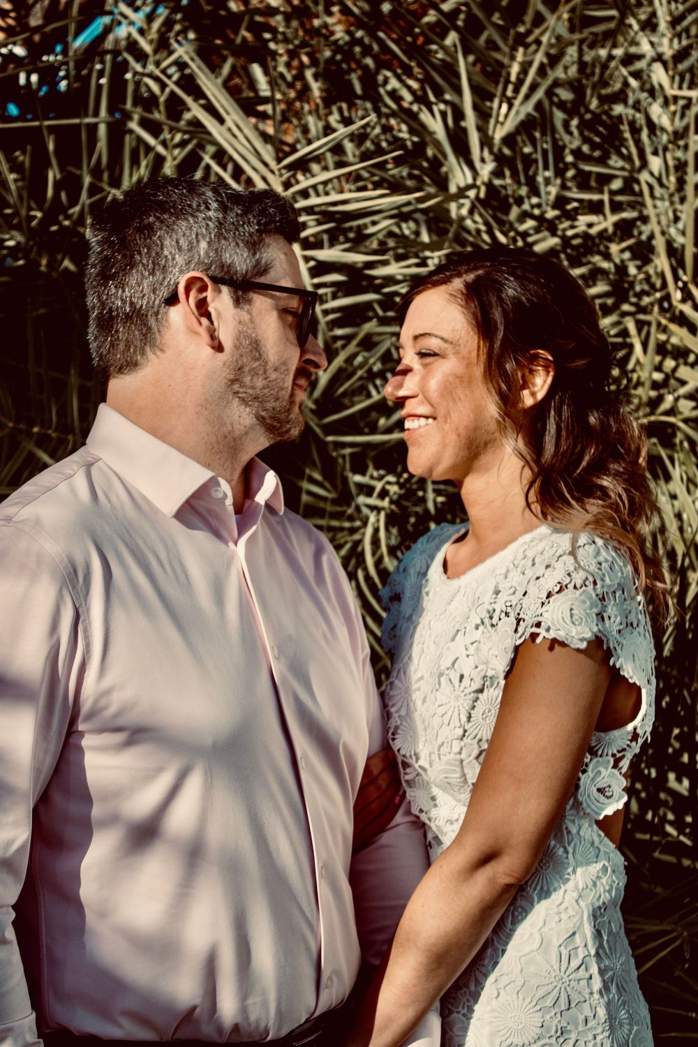 Smiling bride in white lace dress gazes at groom in pink shirt against tropical palm background.