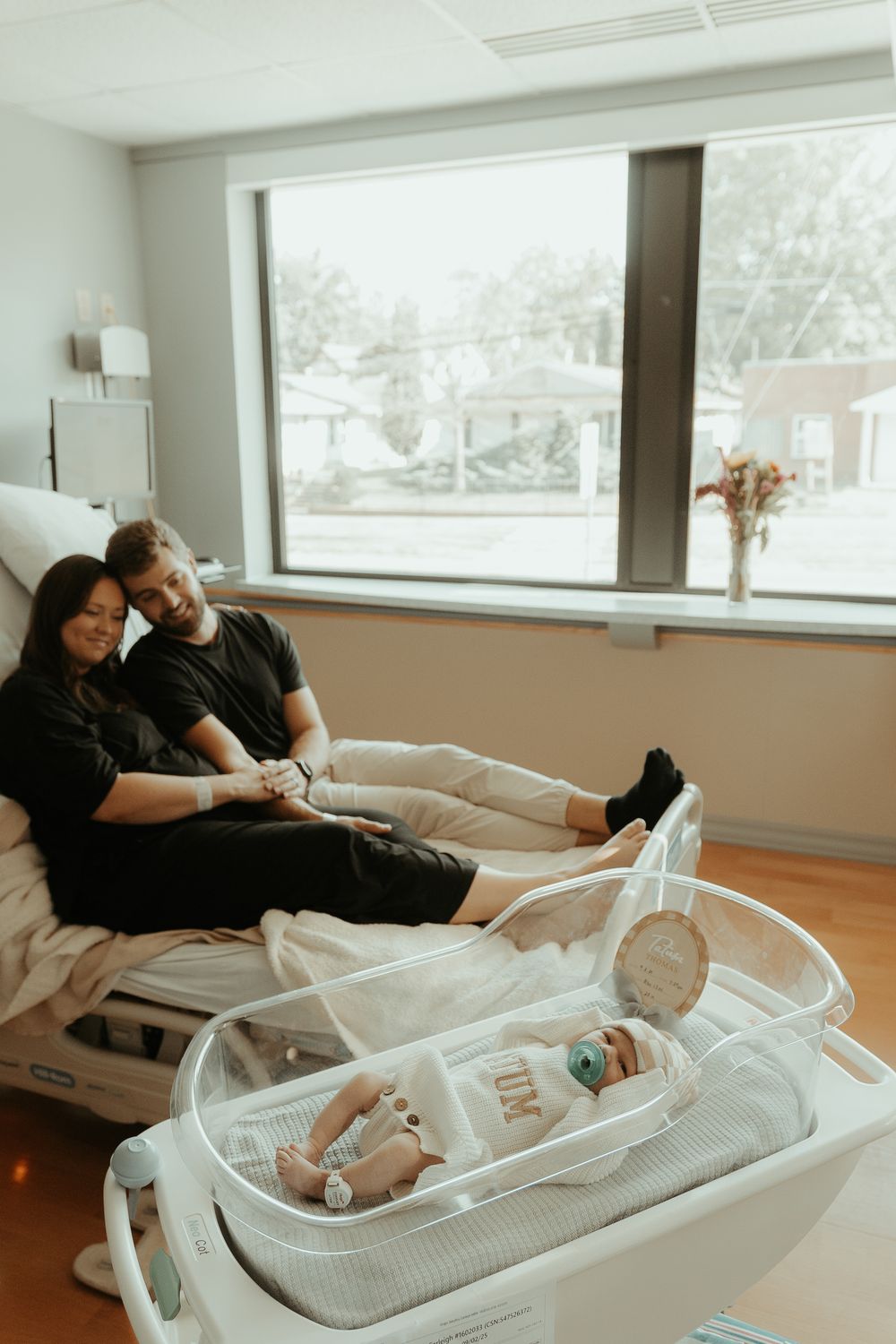 New parents watching over newborn in clear hospital bassinet by window.