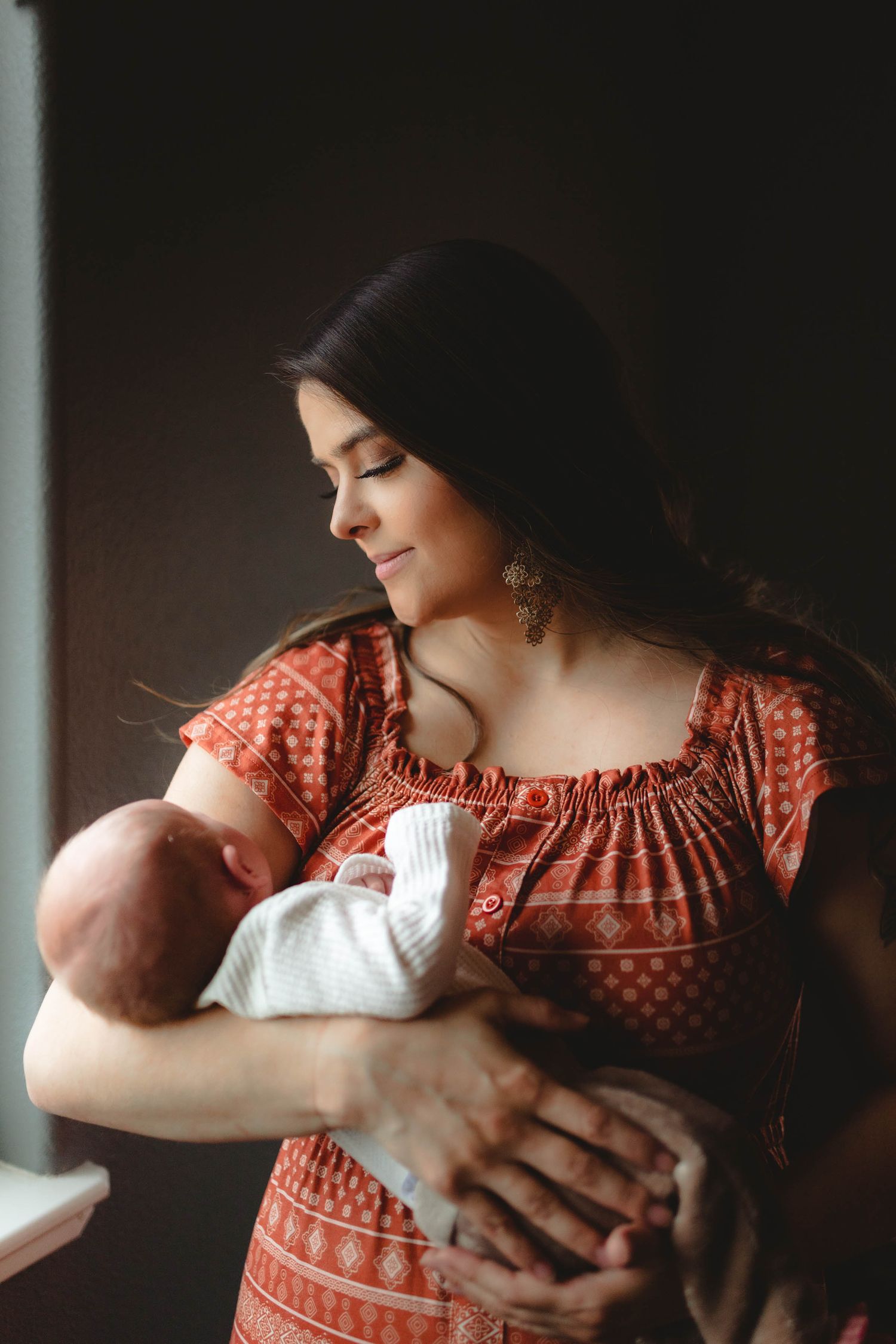 Beautiful dark-haired mother in red patterned dress tenderly holding sleeping newborn by a window.