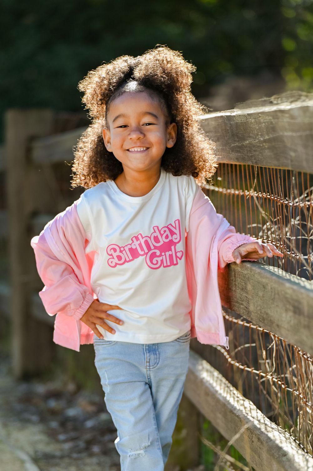 Child wearing pink birthday shirt and jeans poses by wooden fence outdoors.