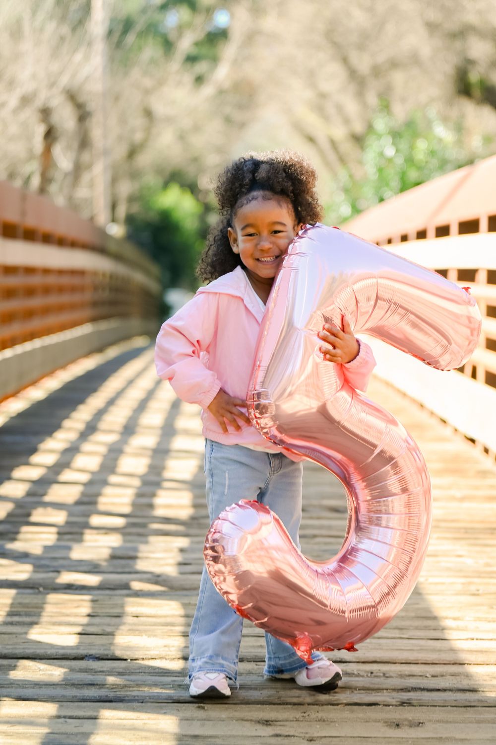 Child holding pink number balloon poses on wooden bridge in casual outfit.