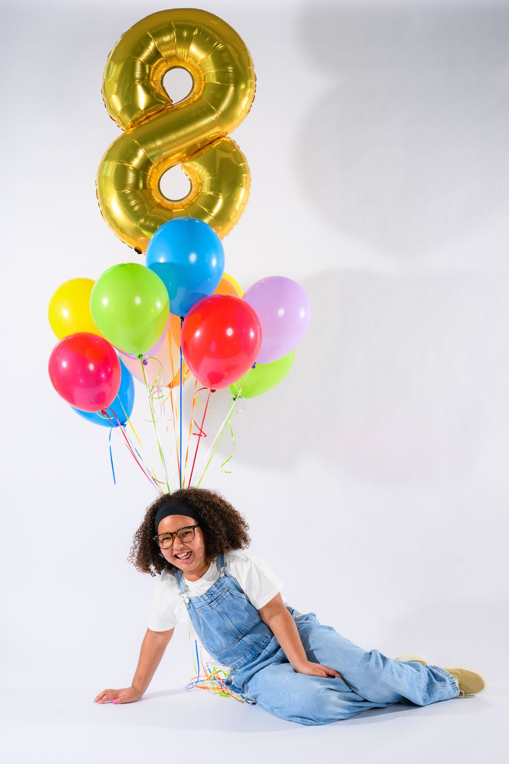 Happy child sitting on floor holding colorful balloons with gold number 8 balloon, wearing denim overalls and glasses.