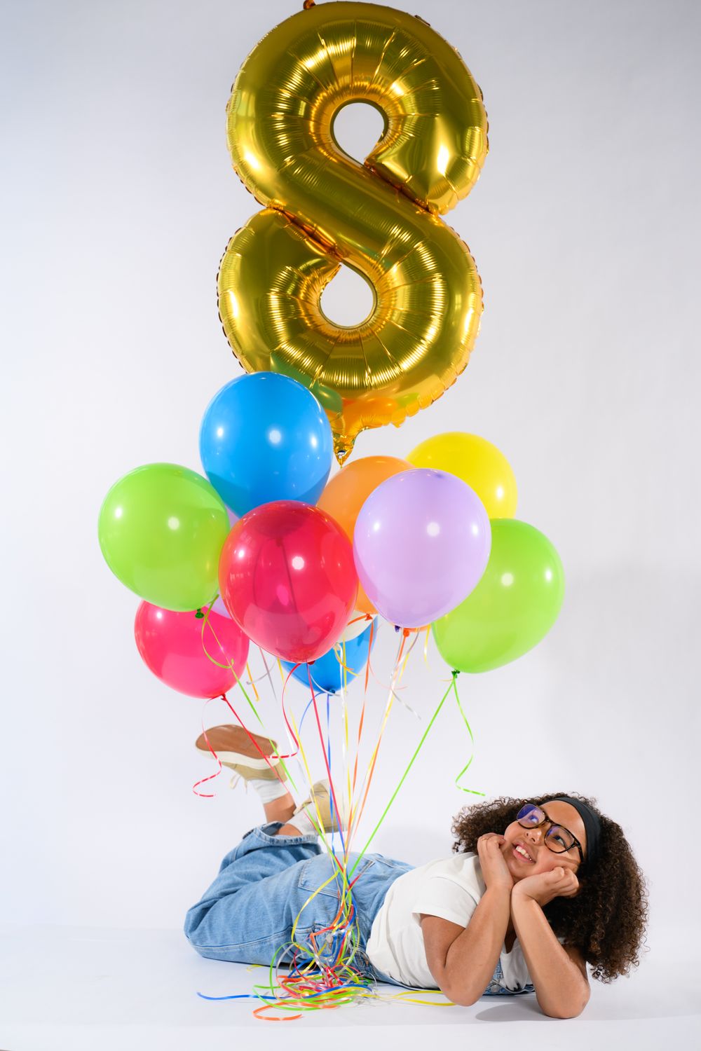 Young girl lying on floor with colorful balloons and gold number 8 balloon on white background.