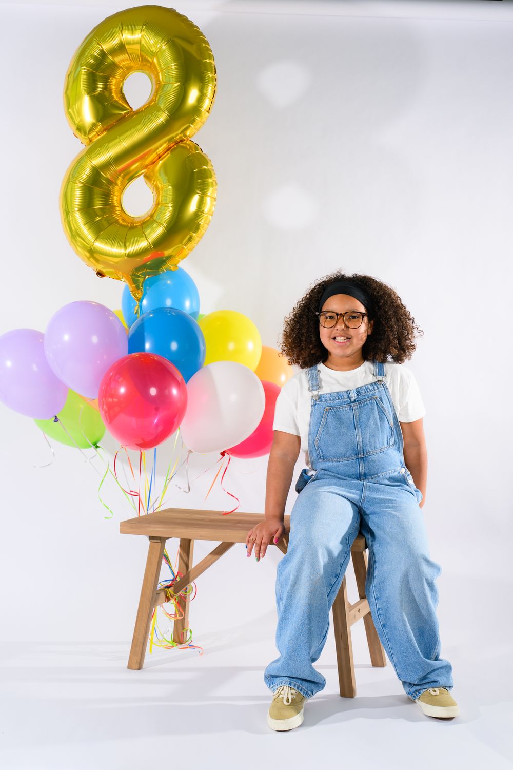 A smiling child in denim overalls sits beside colorful balloons and a gold number 8 balloon on white background.