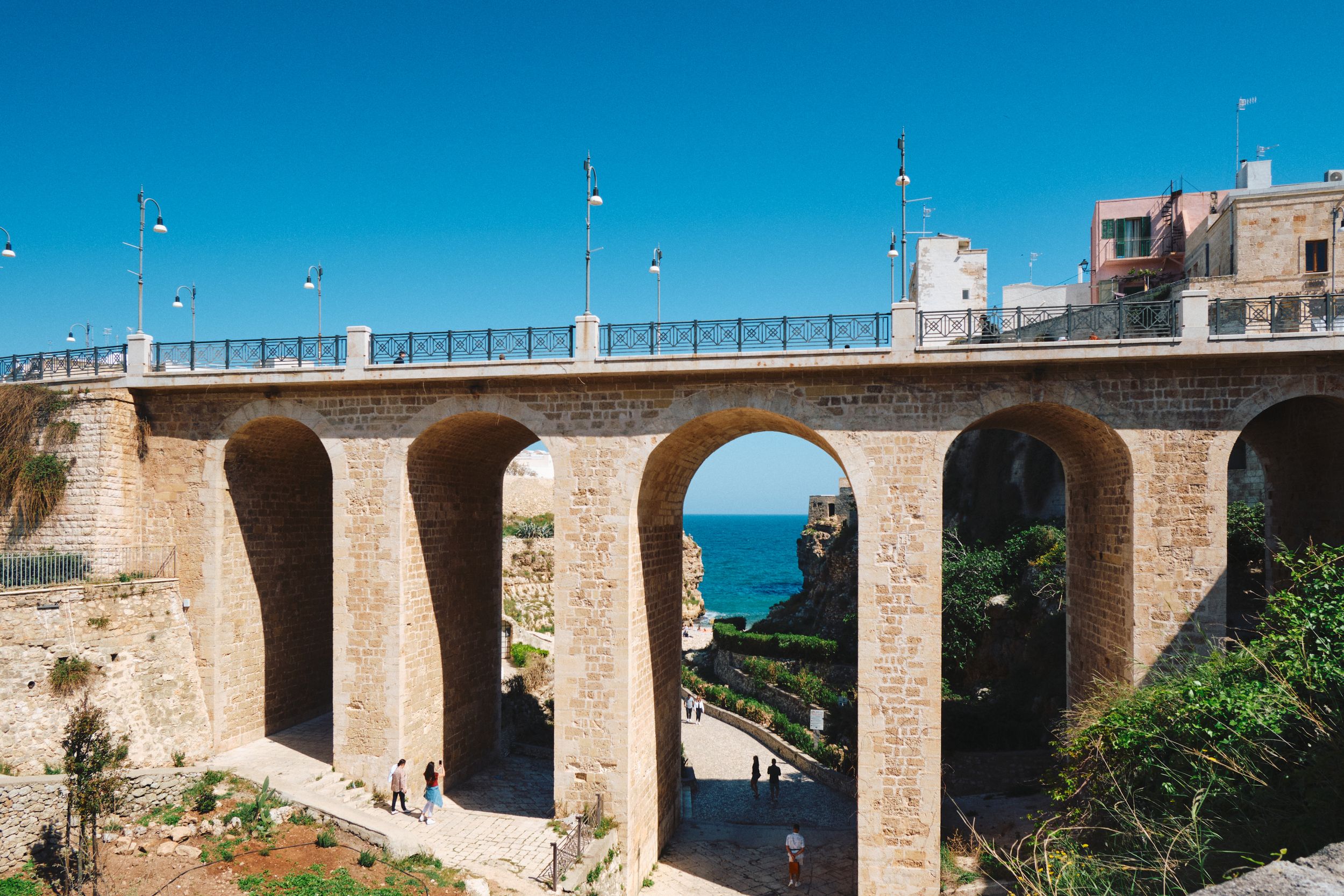 Stone viaduct bridge with arches overlooking the Mediterranean Sea against bright blue sky.