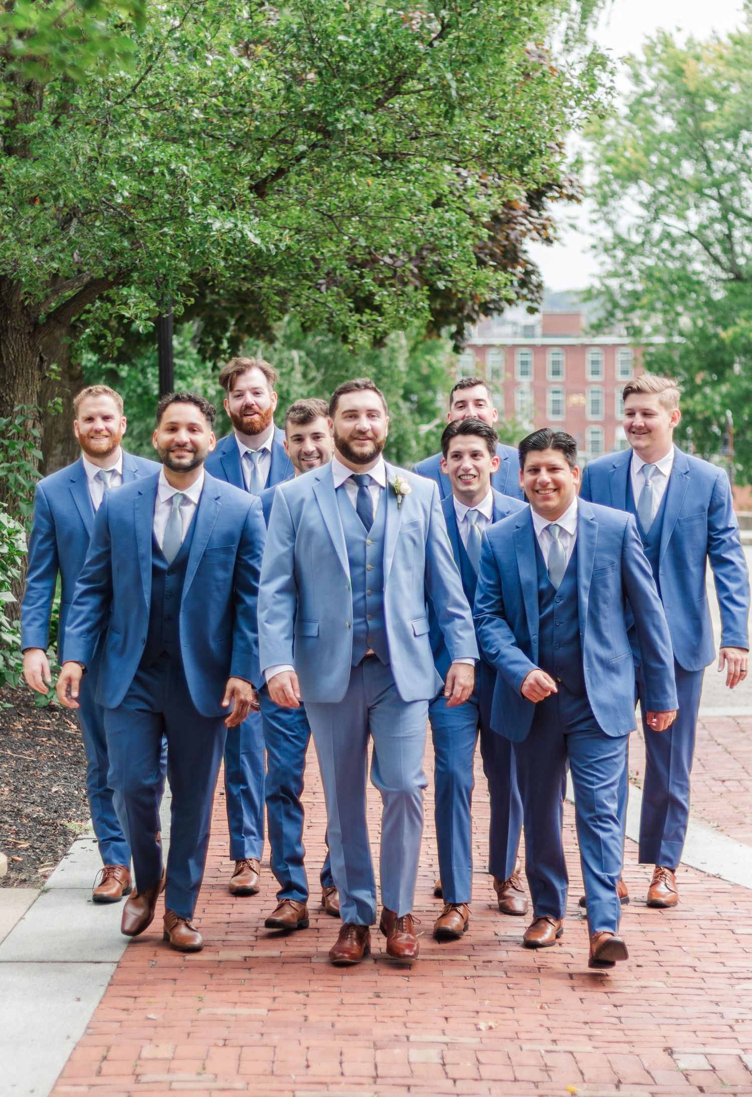 Groomsmen in matching blue suits walk together on brick pathway.
