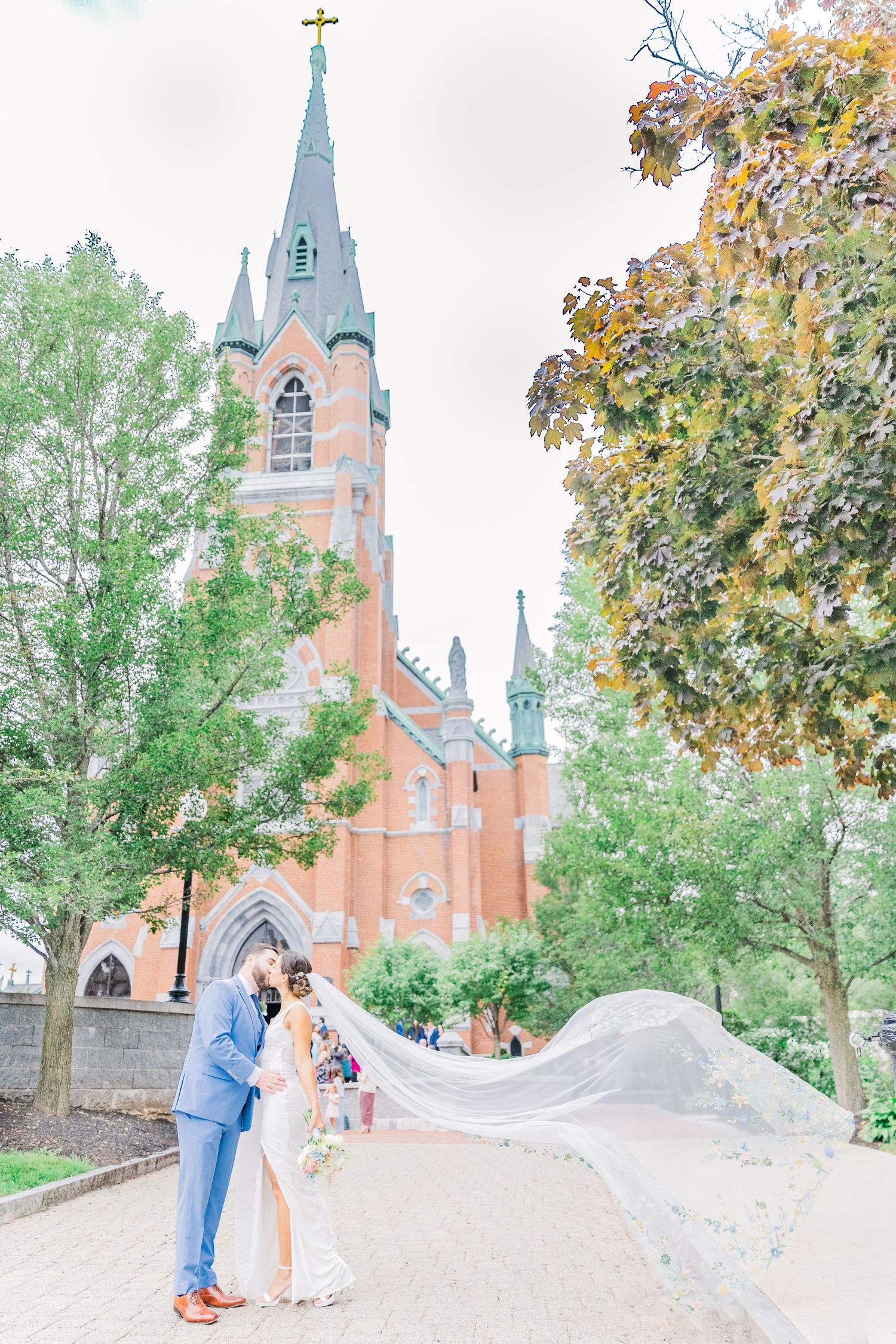 Flowing veil catches breeze as couple poses before historic Gothic church with copper spire.