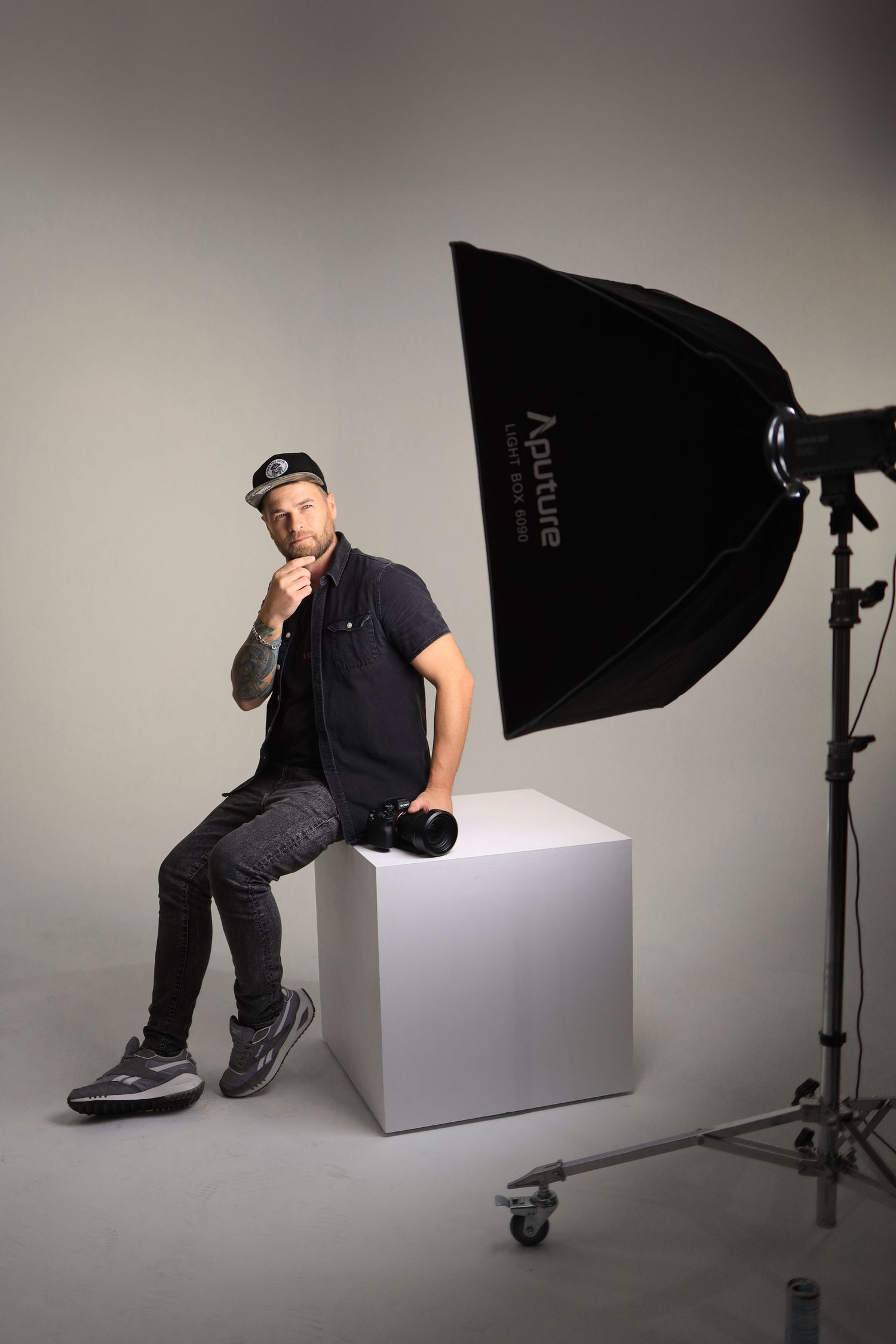 Professional studio portrait of person in casual wear posing on white cube with lighting equipment.