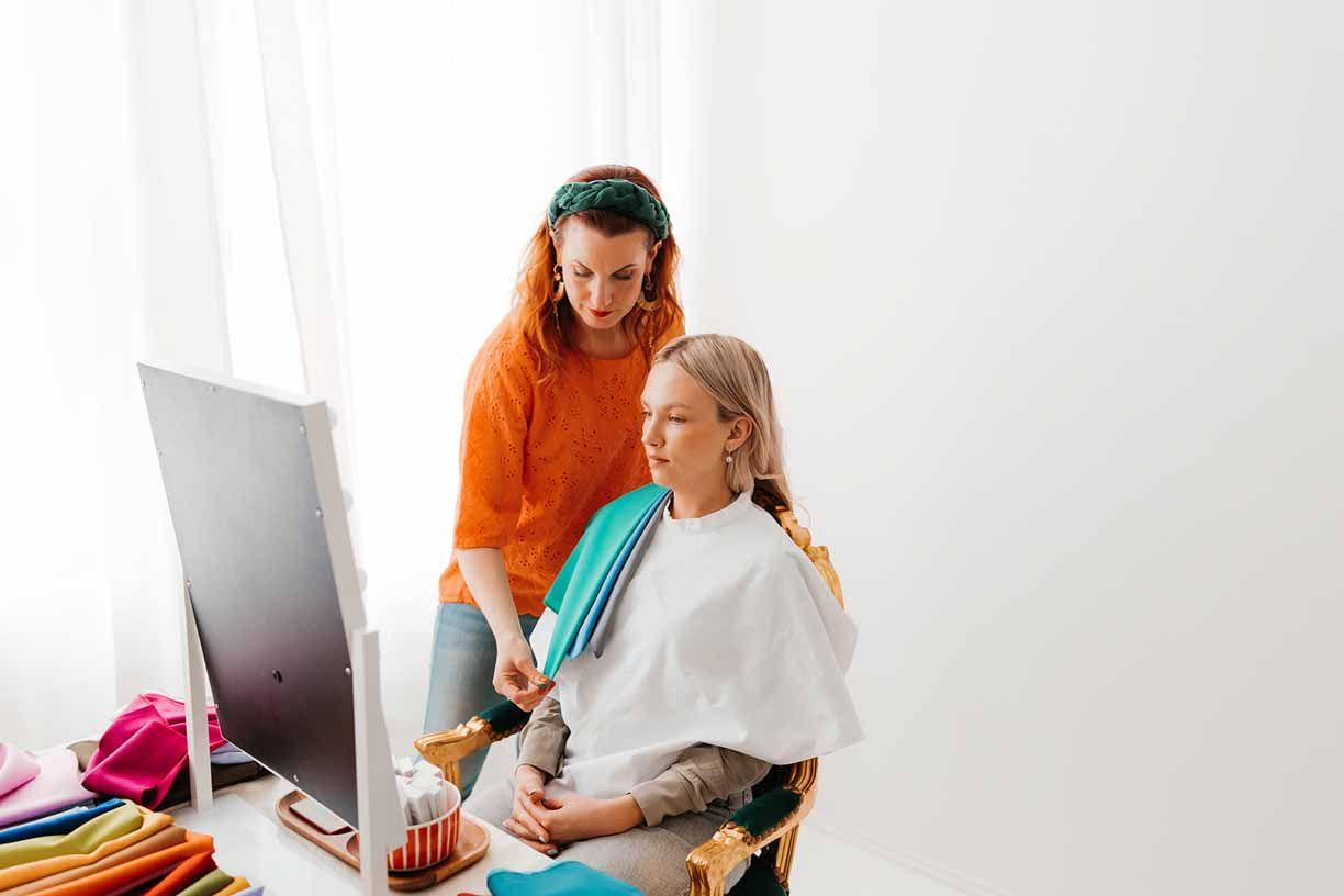 A hairstylist in orange consults with client in turquoise while looking at computer screen.