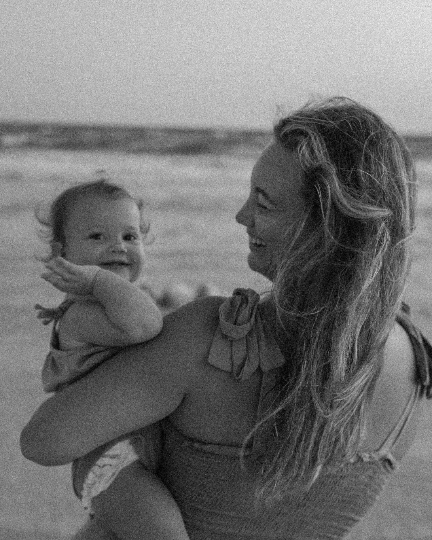 Black and white beach portrait of mother holding baby against ocean backdrop.