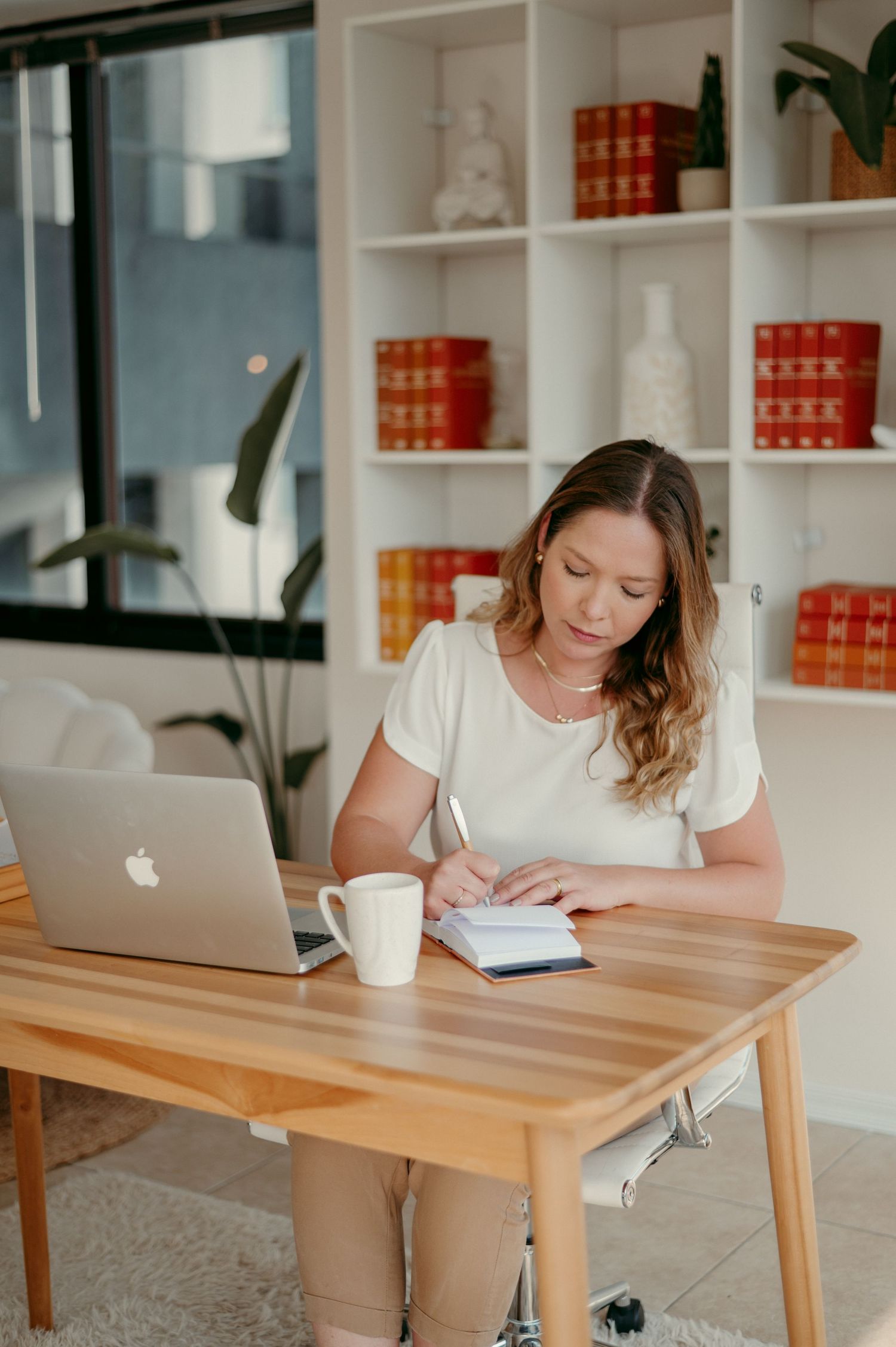 Woman working at wooden desk with MacBook and notes in modern office space.