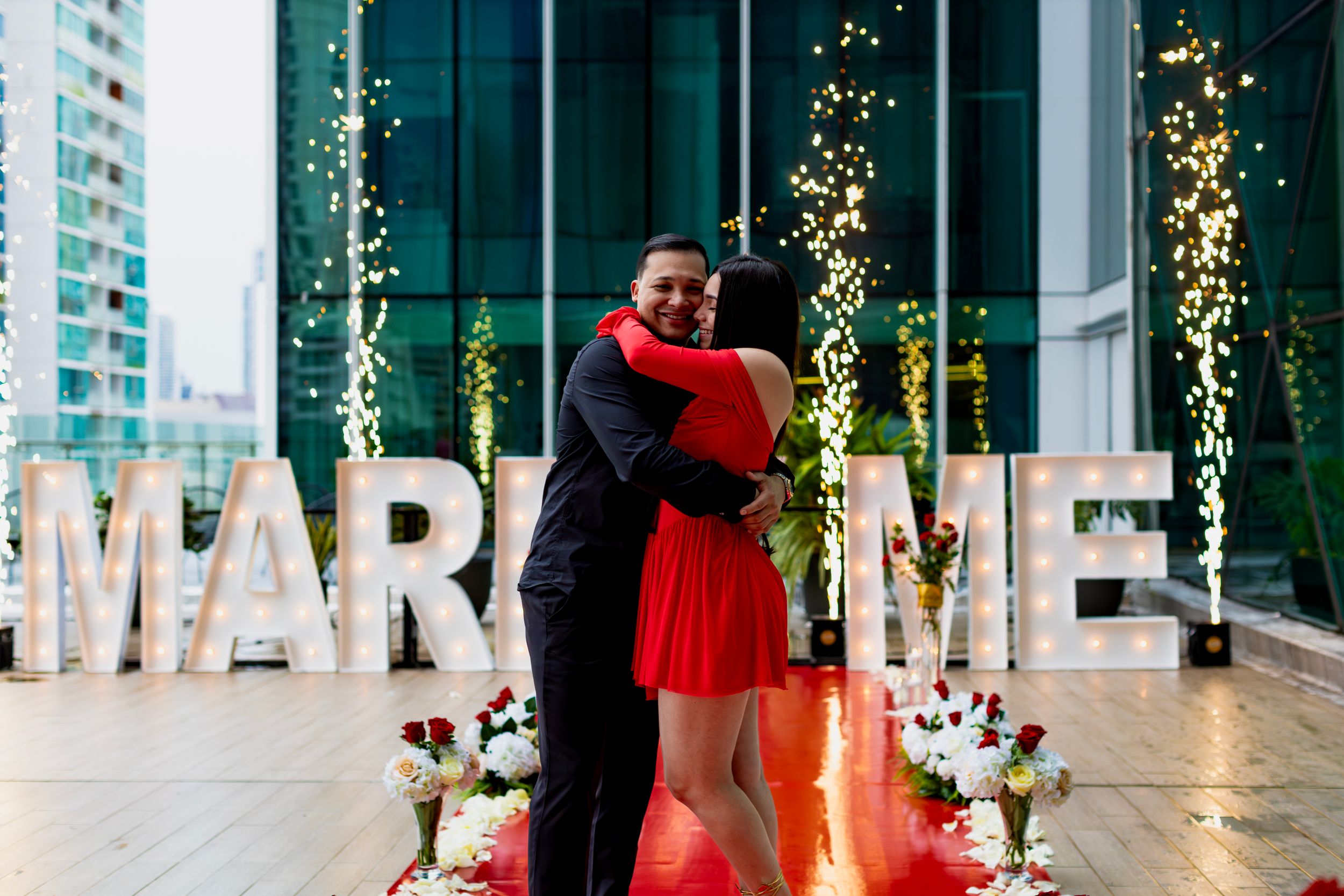 A romantic proposal scene with illuminated 'MARRY ME' letters and hanging lights in the background.