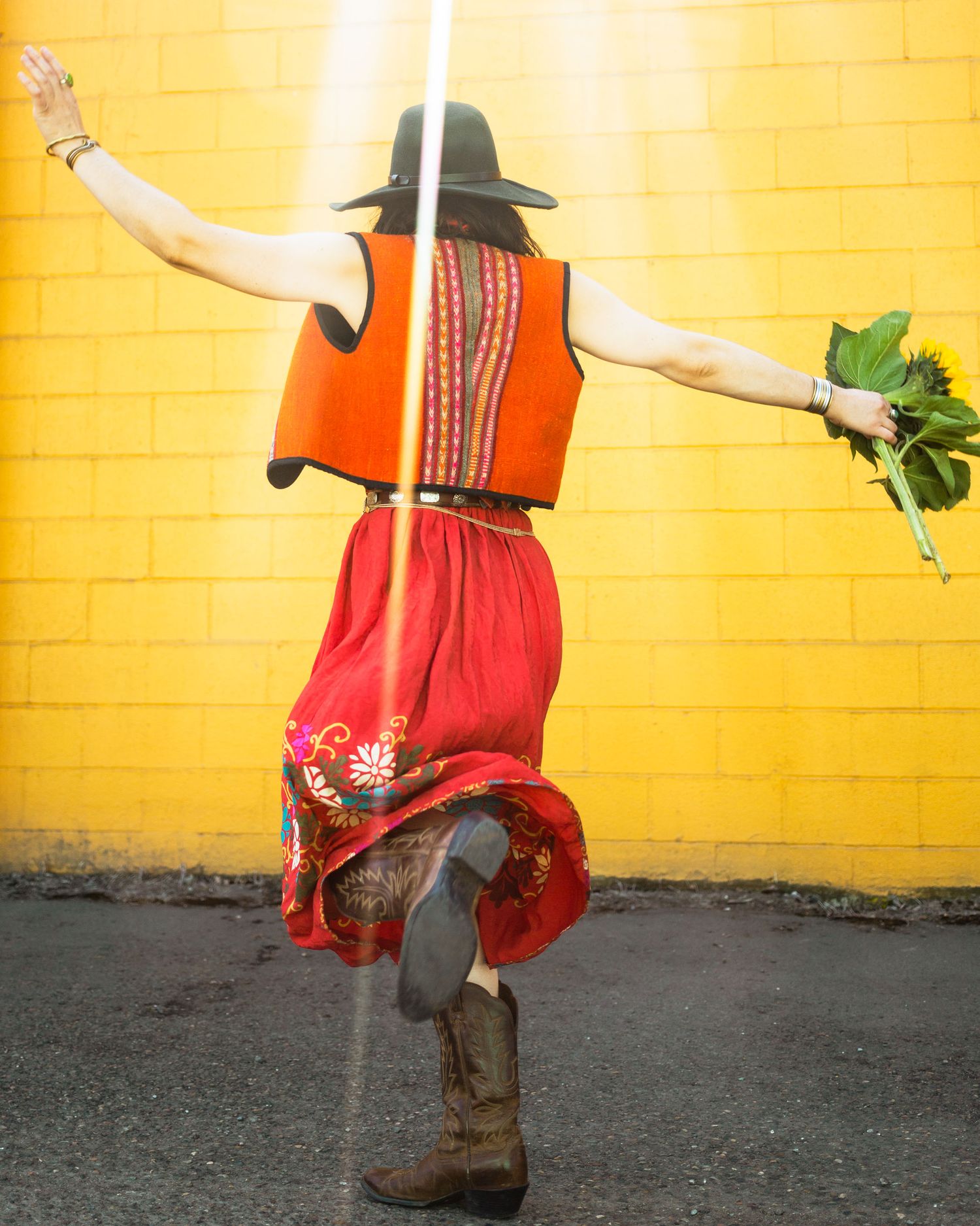Someone in a red skirt and orange vest dances against a bright yellow wall while holding fresh greens.