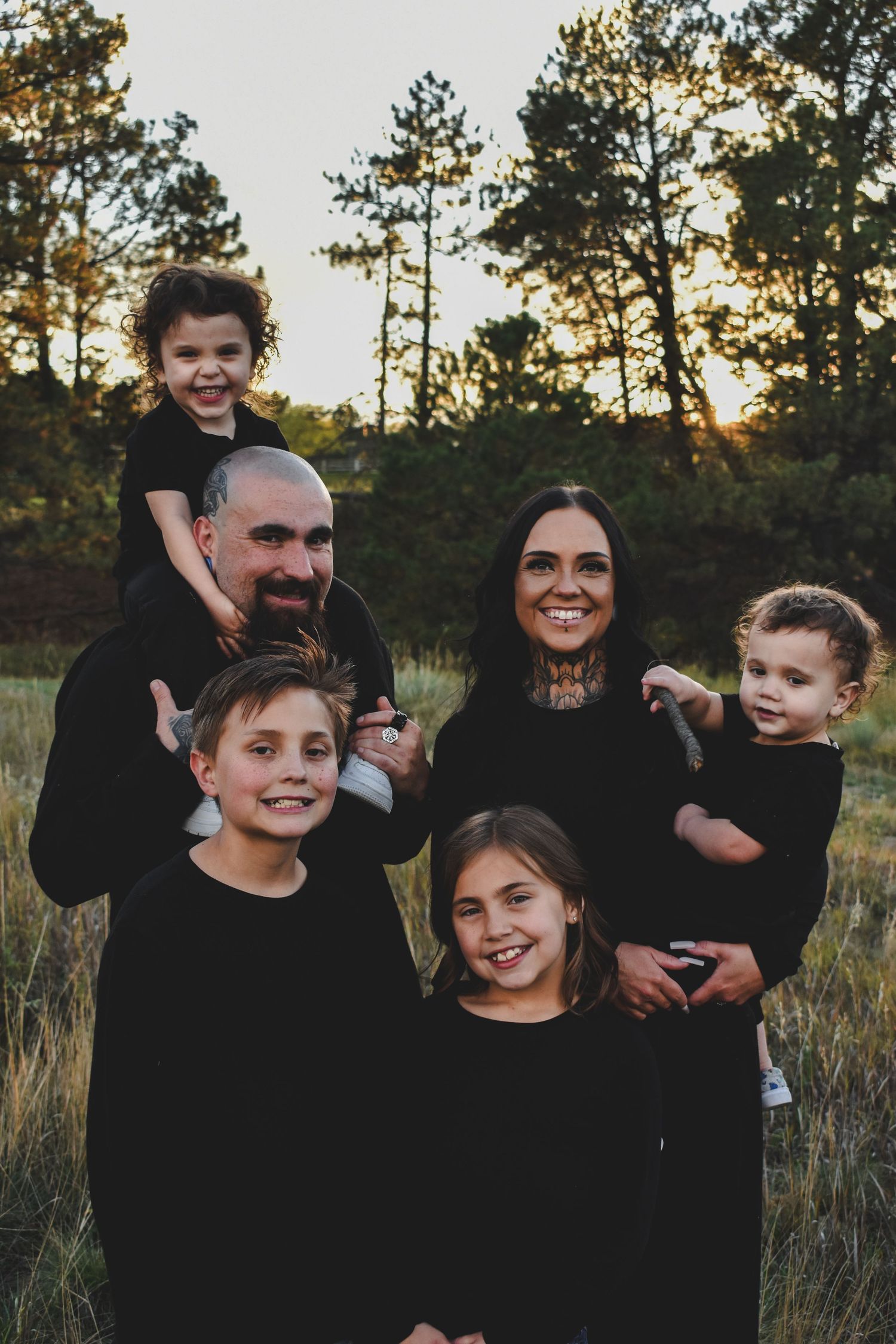 A happy family of six dressed in black poses together in a sunlit forest meadow at golden hour.