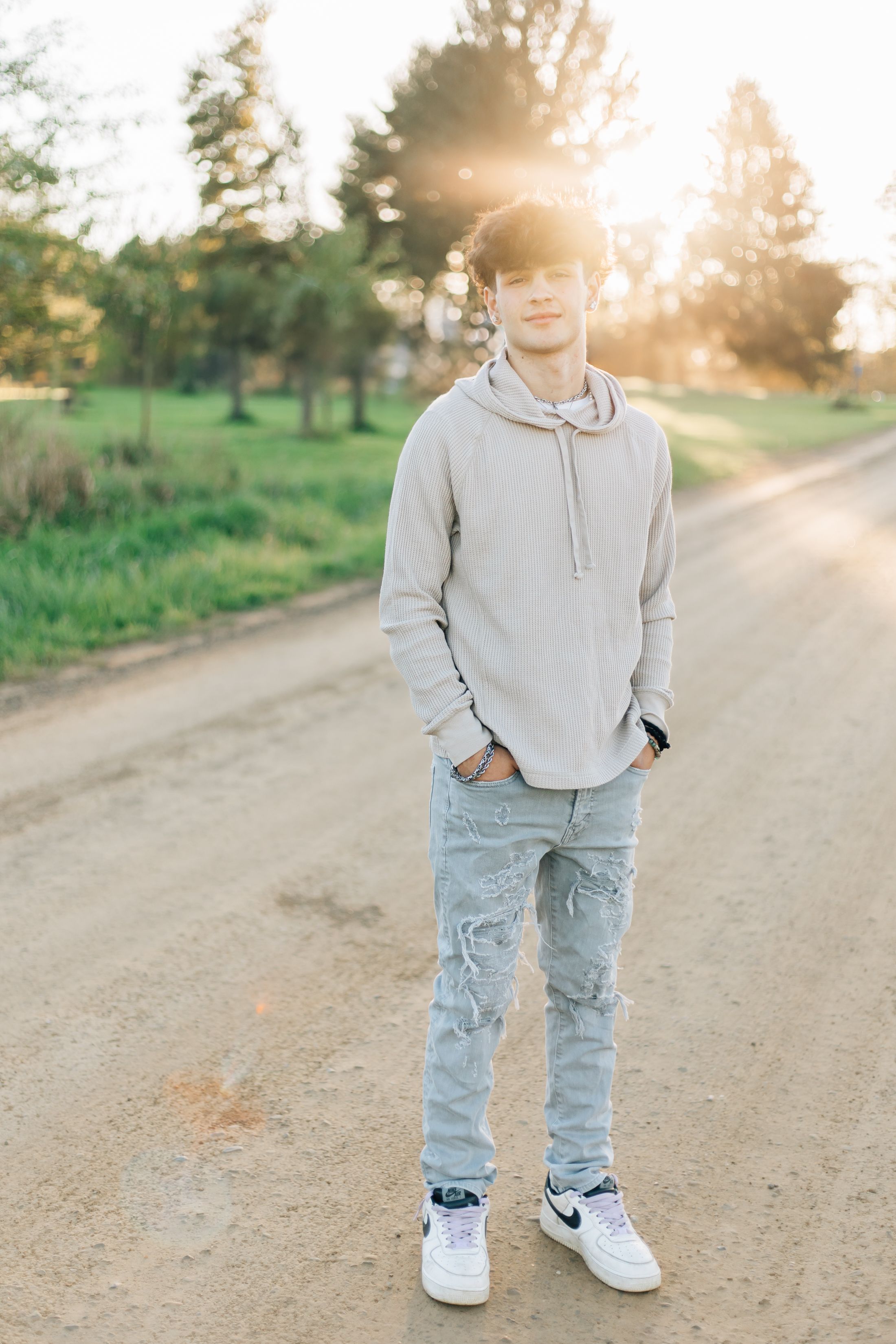 Person wearing casual hoodie and distressed jeans standing on rural path with sunset in background.