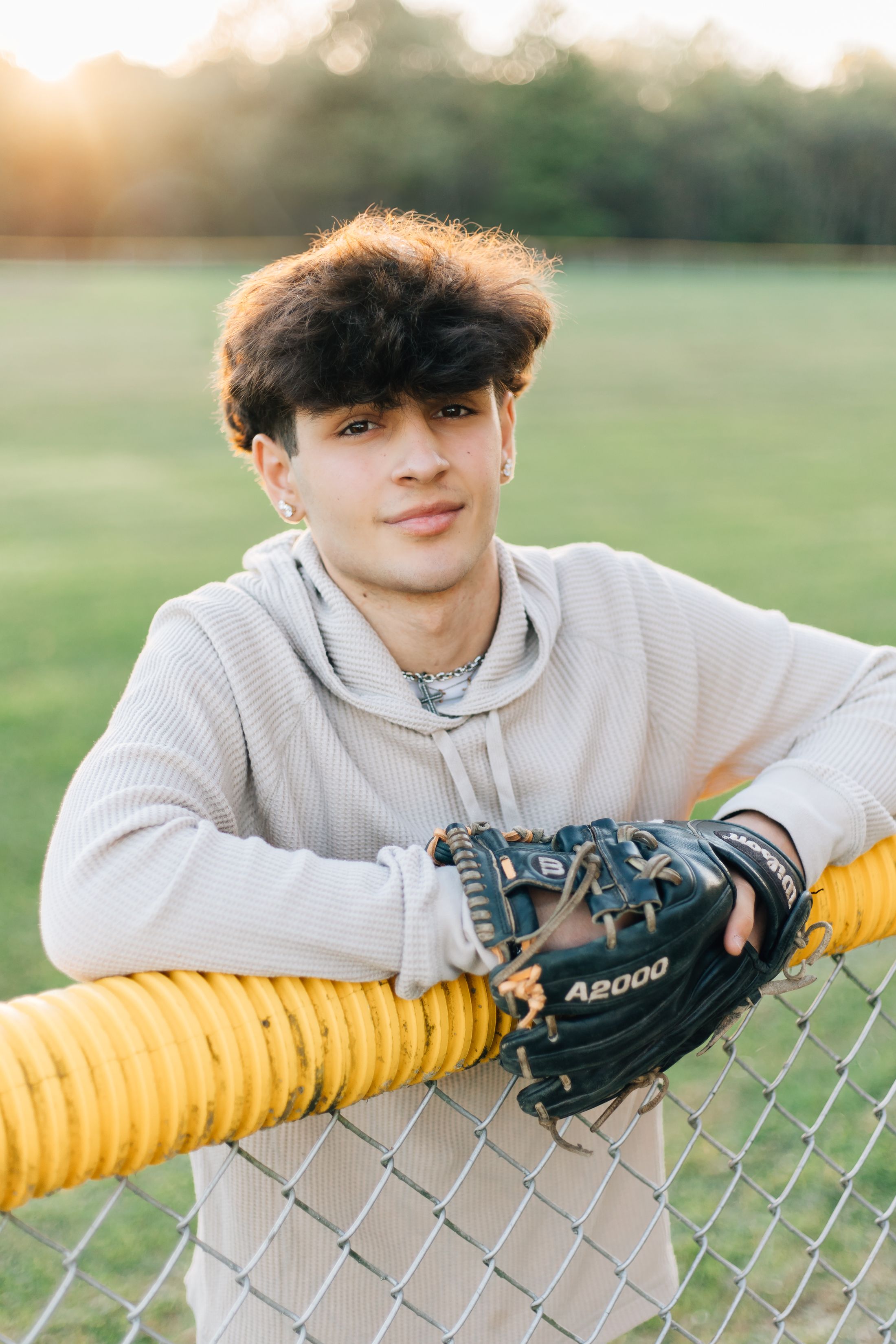 Baseball player in grey hoodie leaning against fence at sunset with glove.