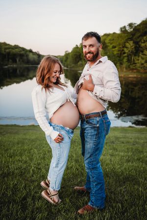 A couple in jeans and white shirts shares tender moments by a lake during a maternity photoshoot at sunset.