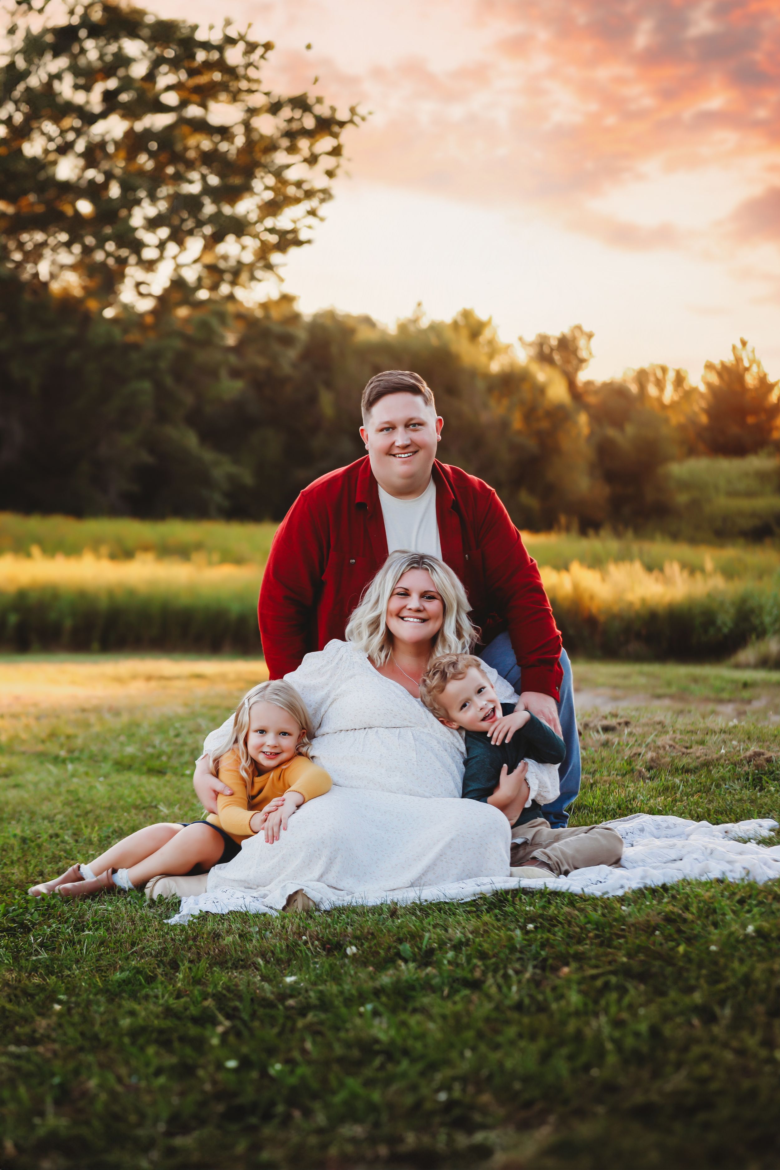 Family sits together on blanket in grassy field during golden sunset with trees in background.