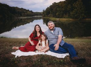 A couple in red and blue outfits relaxes on a blanket near a serene lake during an autumn engagement photoshoot.