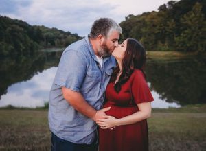 A romantic couple in burgundy and blue outfits share tender moments by a serene lake during an outdoor photoshoot.