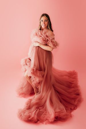 Ethereal maternity portrait featuring a flowing dusty rose tulle gown with ruffled details against a pink backdrop.