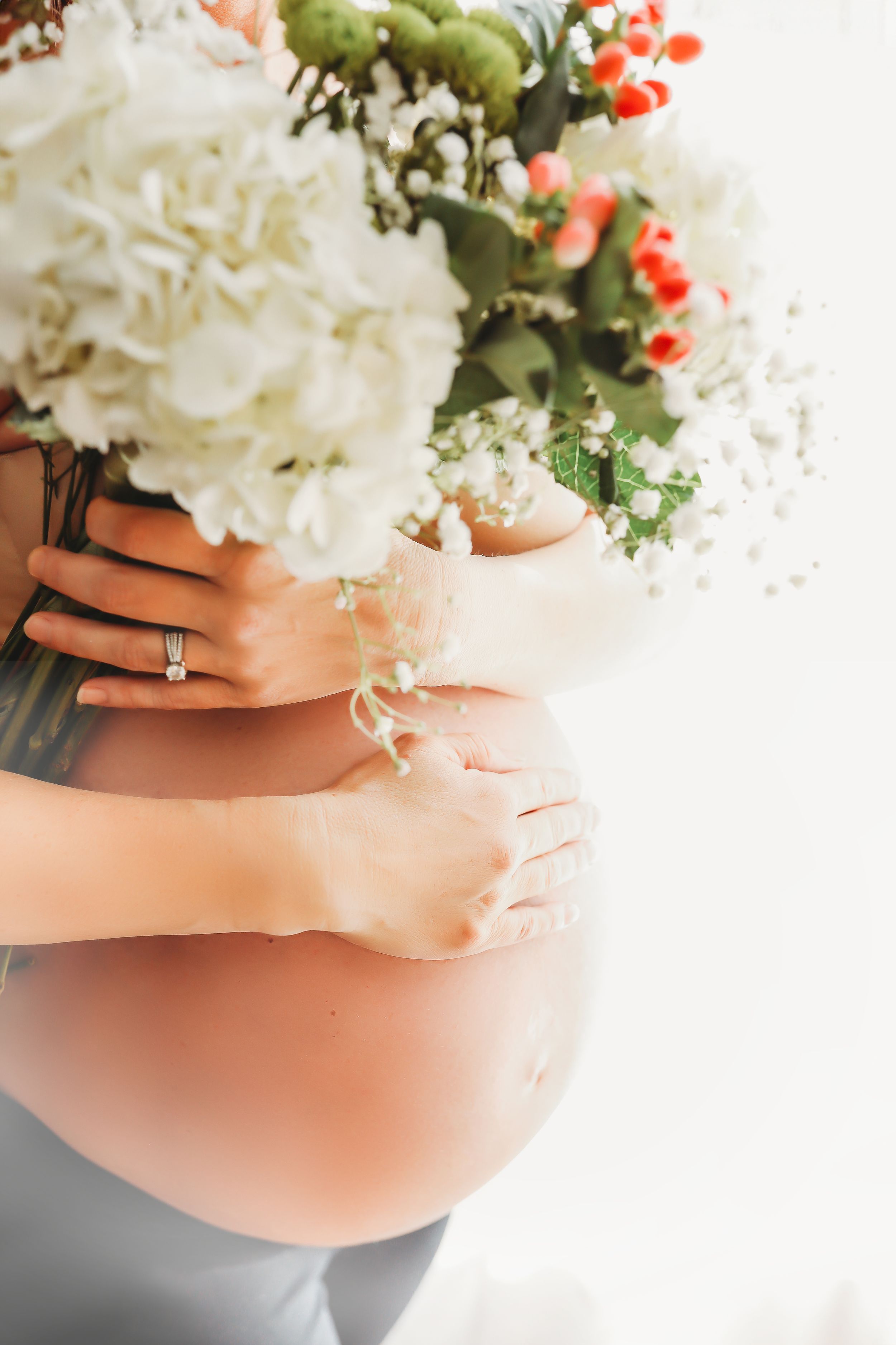 A pregnant woman's belly with white hydrangea flowers and red berries creating a beautiful maternity photo composition.